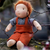 A handmade felt doll with orange hair, wearing a gray shirt and rust-orange overalls, sitting on a tree branch outdoors.