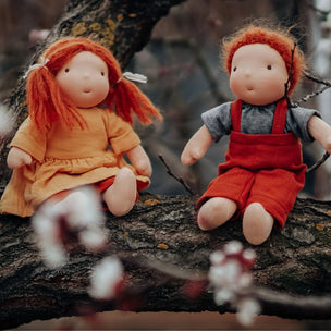 Two handmade wool dolls with red hair sit on a tree branch outdoors. One doll wears an orange dress, and the other wears a gray shirt with red overalls.