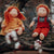 Two handmade wool dolls with red hair sit on a tree branch outdoors. One doll wears an orange dress, and the other wears a gray shirt with red overalls.