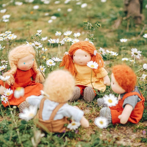 Three soft fabric dolls with curly and wavy hair sit in a grassy field of white daisies. One doll wears an orange dress, another wears a yellow dress, and the third wears orange overalls over a gray shirt.