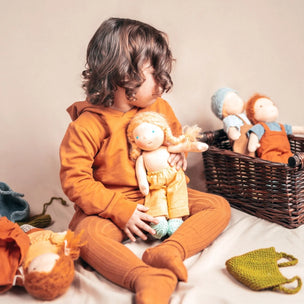 A child with curly brown hair sits on a white blanket, wearing a matching burnt orange long-sleeve top and leggings and holding a handmade doll. A wicker basket filled with more dolls and a green knitted pouch are visible in the background against a warm beige wall.