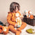 A child with curly brown hair sits on a white blanket, wearing a matching burnt orange long-sleeve top and leggings and holding a handmade doll. A wicker basket filled with more dolls and a green knitted pouch are visible in the background against a warm beige wall.