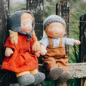 Two handmade paper dolls sitting on a rustic wooden fence outdoors. One doll wears a red dress and gray bonnet with yellow hair, and the other wears brown overalls with a gray bonnet and white shirt.