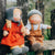 Two handmade paper dolls sitting on a rustic wooden fence outdoors. One doll wears a red dress and gray bonnet with yellow hair, and the other wears brown overalls with a gray bonnet and white shirt.