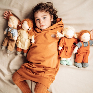 A child lying on a beige bed wearing a matching rust-orange hooded sweatshirt and ribbed knit pants, with four soft cloth dolls arranged beside them.