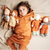 A child lying on a beige bed wearing a matching rust-orange hooded sweatshirt and ribbed knit pants, with four soft cloth dolls arranged beside them.