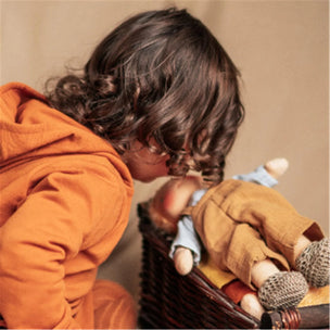 A child in a warm orange hoodie leans over a basket containing a small doll dressed in tan and light blue, against a soft beige background.