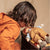 A child in a warm orange hoodie leans over a basket containing a small doll dressed in tan and light blue, against a soft beige background.