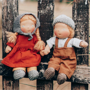 Two handmade cloth dolls sitting side by side on a rustic wooden fence. The doll on the left has long blonde braids, a grey knit bonnet, and a rust-red dress.