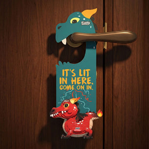 A decorative door hanger shaped like a dragon mouth hanging on a wooden door. The teal dragon head is hung on a brass-colored doorknob, and has the text 'IT'S LIT IN HERE.