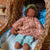 A handmade African American doll with curly dark hair, wearing a floral peach top and light green pants, resting inside a woven wicker bassinet with a blue patterned lining.