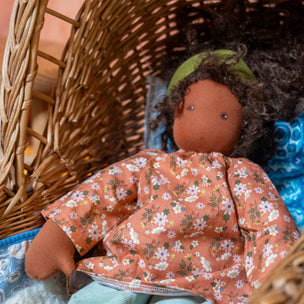 A handmade cloth doll with dark brown skin, curly dark hair, and a green headband, wearing a peach floral long-sleeve top and mint green pants, resting inside a woven wicker basket.