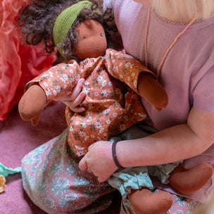 A young child with blonde hair sits on a pink carpet, holding and kissing a handmade cloth doll with curly brown hair, a green headband, and a floral dress.