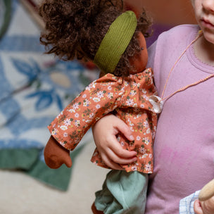 A child in a pink shirt holds a doll with curly brown hair, wearing a green headband, a rust-orange floral long-sleeve top, and sage green pants.