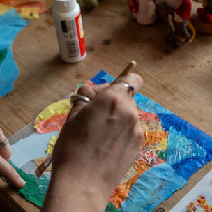 Person painting a colorful piece of art on a wooden table with glue and other items.