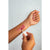 A close-up of a person's forearm with a mauve-pink lip gloss swatch applied, while another hand holds the applicator above the swatch. The product tube is white with a pink cap, shown against a light gray background.