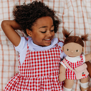 Child in a red checkered dress lying on a checkered blanket with a doll wearing a matching outfit.