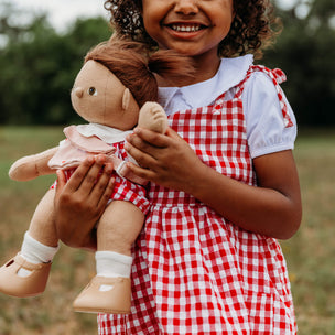 Child in a red and white checkered dress holding a doll outdoors.