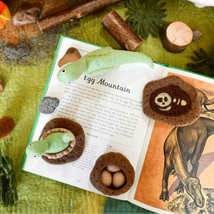 Open book with illustrations of eggs and a dinosaur, surrounded by wooden logs and a toy dinosaur on a green surface.