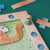 A close-up of a wooden children's board game on a dark green surface. The game features a light wood path with blue numbered holes, a green illustrated play mat with a winding tan track and a small house, and small wooden pieces including blue crosses and tan slats.