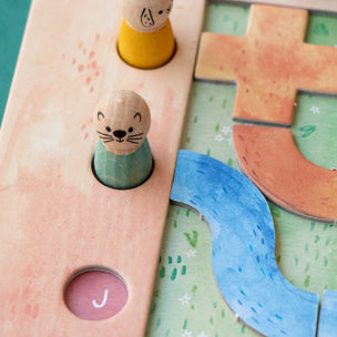 Close-up of a wooden children's puzzle board with colorful pieces, including a blue winding river, tan letter-like shapes, and small animal pegs on a peach-colored track.