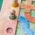 Close-up of a wooden children's puzzle board with colorful pieces, including a blue winding river, tan letter-like shapes, and small animal pegs on a peach-colored track.