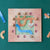 A square wooden puzzle board with a pastel landscape design, surrounded by scattered wooden puzzle pieces on a dark green background.