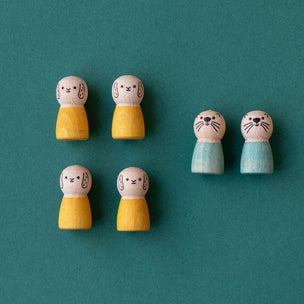 Five small wooden toy figures with cat faces are arranged on a teal background. Four figures are yellow, and two are light blue.