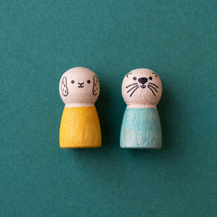 Two small wooden toy figures with animal faces, one wearing a yellow outfit and the other wearing a light blue outfit, standing on a teal background.