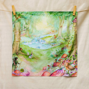 A square fabric art print or tea towel featuring a whimsical watercolor forest scene with a pond, trees, flowers, and small animals, hanging from a string with wooden clothespins on a cream fabric background.