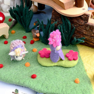 A miniature felt forest play scene with a purple and pink felted animal on a green hill, a small white felted creature, dark green felt plants, scattered red and orange felt berries, and a wooden staircase in the background.