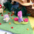 A miniature felt forest play scene with a purple and pink felted animal on a green hill, a small white felted creature, dark green felt plants, scattered red and orange felt berries, and a wooden staircase in the background.
