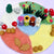A circular felt play rug depicting a seasonal landscape with four small felted character figures standing in the center, surrounded by mushrooms, leaves, grass, and colored sections representing seasons.