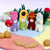A colorful felt garden scene with small mushroom-like figures in purple, green, red, and white, a red mushroom, tan acorns, and green leaves on a pastel round mat against a light lavender background.