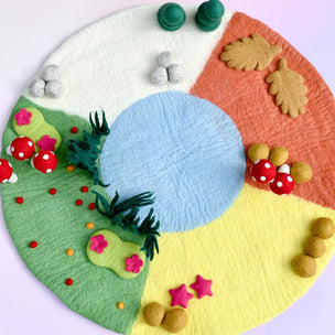 A circular felt craft rug or play mat featuring four colored quadrants: white, orange, yellow, and green, with small felt trees, mushrooms, flowers, leaves, and stars attached around the border.