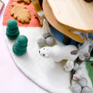 A close-up of a handmade felted wool playset featuring a white polar bear, green felted trees, gray felted rocks, and a light wood platform on a pastel pink and white background.