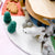 A close-up of a handmade felted wool playset featuring a white polar bear, green felted trees, gray felted rocks, and a light wood platform on a pastel pink and white background.