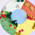 A circular felt craft decoration featuring four colored sections in white, orange, yellow, and green, with a central light blue circle. Decorative elements include felt grass, pink and red flowers, brown leaves, and red-and-white mushrooms, arranged on a pale pink background.