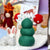 A close-up of a felted wool winter scene with a green tree, red-and-white mushrooms, a white rabbit, and a small white figure with a blue snowflake on its hat, set against a softly blurred forest-themed background.