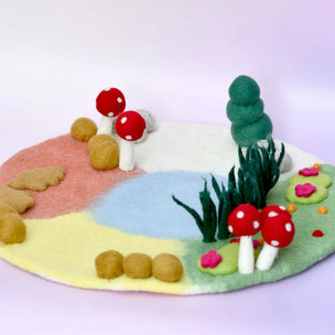 A miniature felt forest scene on a round base, featuring red-and-white mushrooms, green grass, a small green tree, pink flowers, tan stones, and pastel ground sections on a light purple background.
