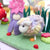 Handmade felt wool art depicting a small pastel-colored sheep beside a purple flower with a cream face, set on a green felt ground with colorful accents.