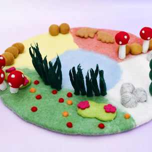 A colorful felt garden play mat with red and white mushrooms, green grass, pink flowers, and a pastel background of yellow, pink, blue, and white sections on a light purple surface.