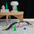 Toy car track set with cars and small trees on a gray surface, with a wooden table and mushroom-shaped object in the background.