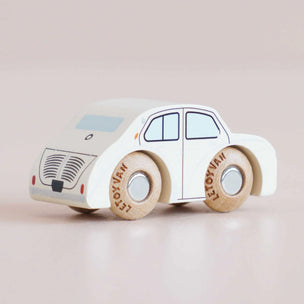 A small wooden toy car with a cream-colored body and natural wood wheels, photographed on a soft beige background. The wheels are printed with the brand name 'LE TOYOVAM'.