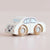 A small wooden toy car with a cream-colored body and natural wood wheels, photographed on a soft beige background. The wheels are printed with the brand name 'LE TOYOVAM'.