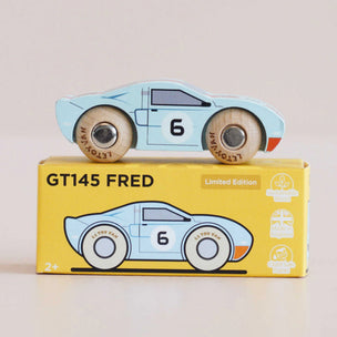 A light blue wooden toy race car with the number 6 sits on top of its yellow 'GT145 FRED' packaging box. The box features an illustration of the same car and small eco-style icons on the right side.
