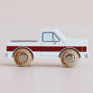 A wooden toy pickup truck with a white body, dark red stripe, and natural wood wheels, shown in profile against a solid pale pink background.