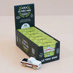 A display box of Le Toy Van 'Charlie' wooden car, showing multiple green individually wrapped wooden toy cars inside, with one white and red wooden car placed in front on a light beige background.