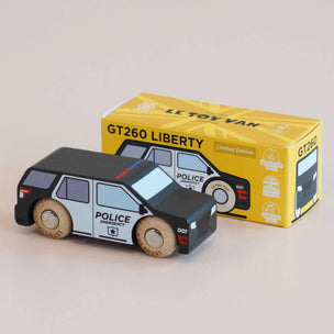 A wooden toy van shaped like a police car, shown in front of its yellow retail box. The toy van is black and white with light wood wheels and small red and blue stripe accents.