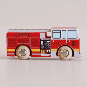 A wooden toy fire engine with a red body, white cab and windows, yellow stripes, and light wood wheels, shown on a plain light beige background.